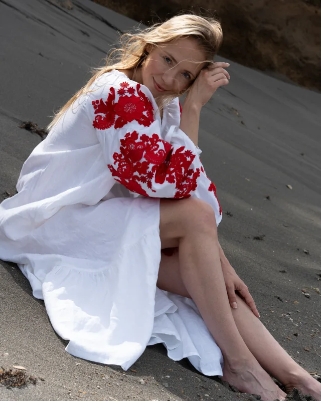 Woman on a beach wearing a white dress with red embroidery, with her hair in the breeze