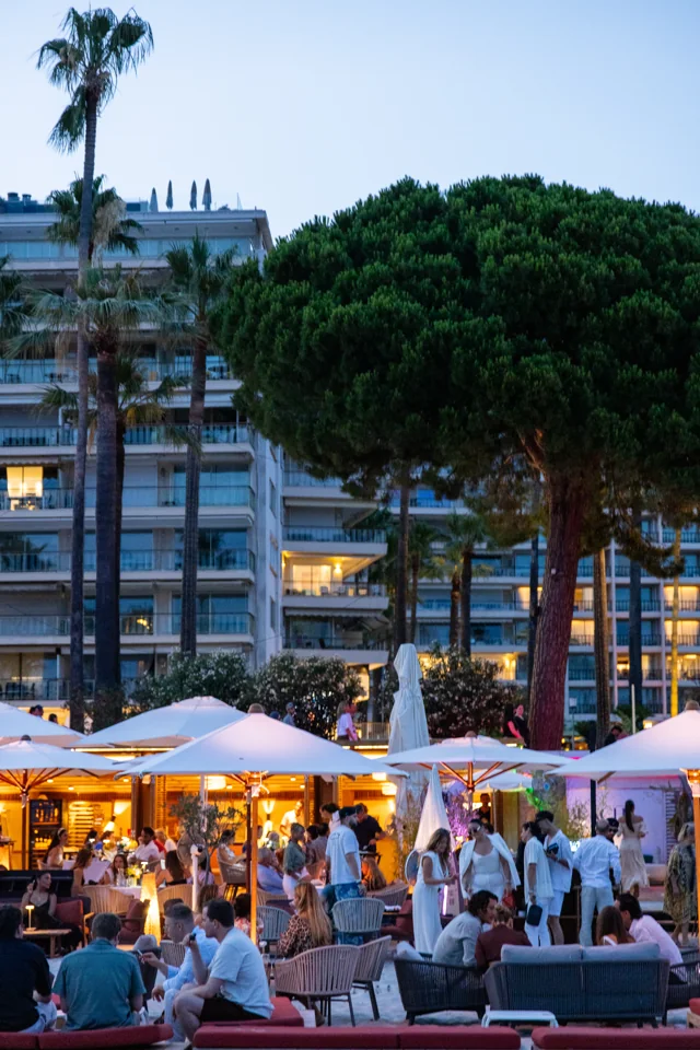 Guests at a beachside reception beneath umbrellas, trees, and illuminated buildings at dusk