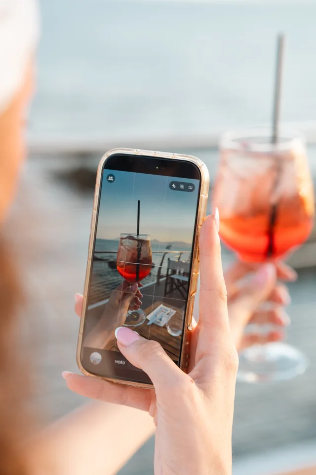 A person photographing a red cocktail with a phone beside the sea
