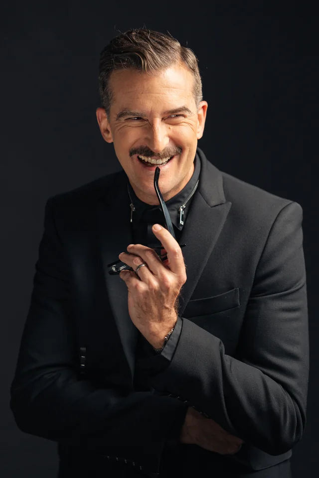 Smiling man in a black suit holding his glasses against a dark studio background