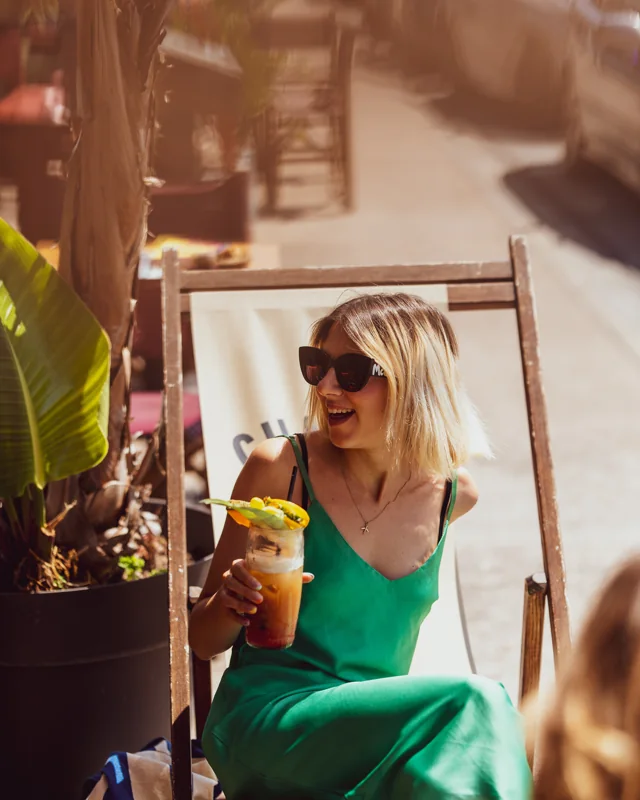 A woman in a green dress laughing with a drink in a sunlit deckchair
